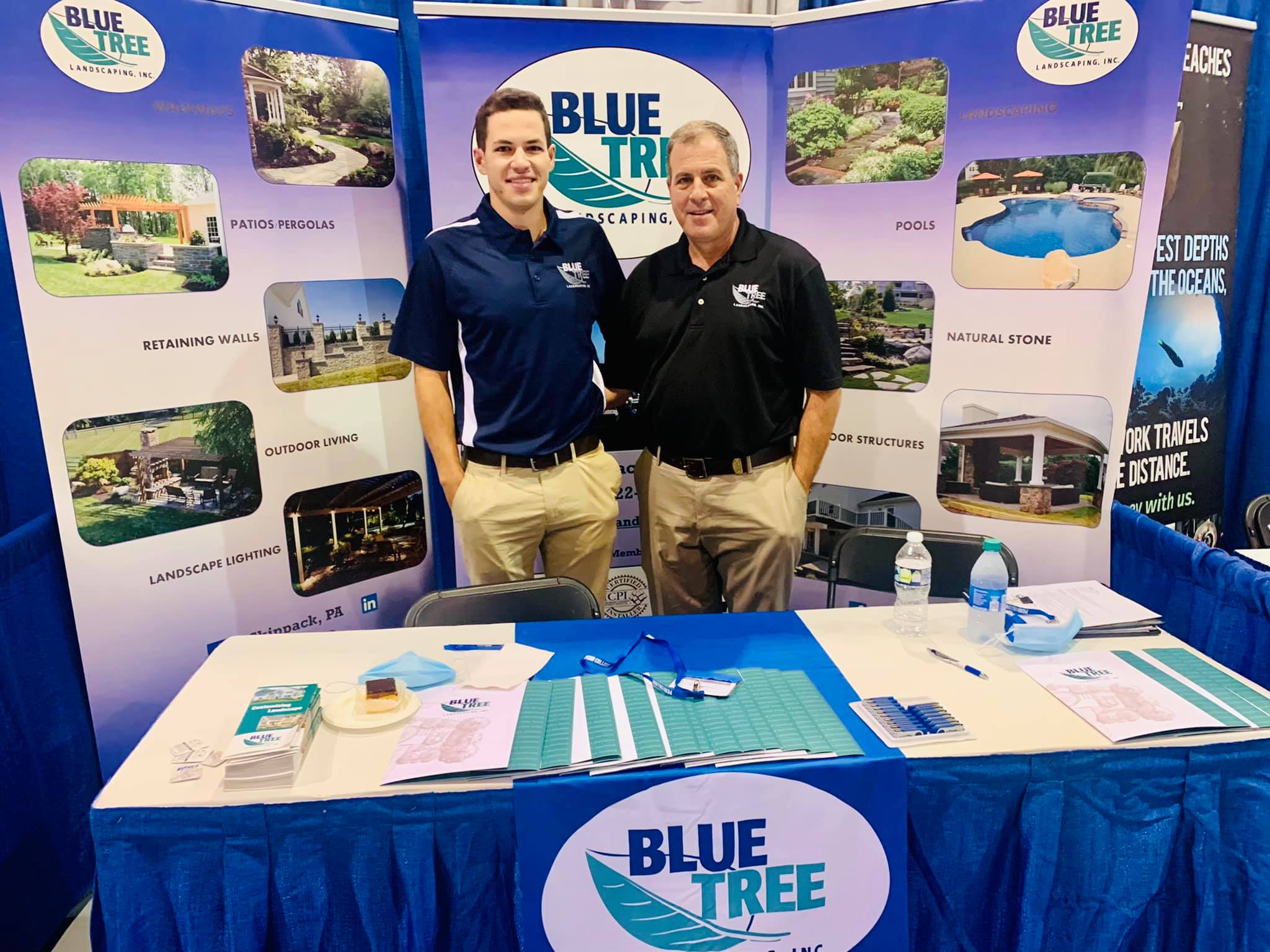 Two men stand smiling at a Blue Tree Landscaping expo booth, which displays banners with images of landscaping projects. The table in front has brochures, business cards, pens, and a bottle of water.