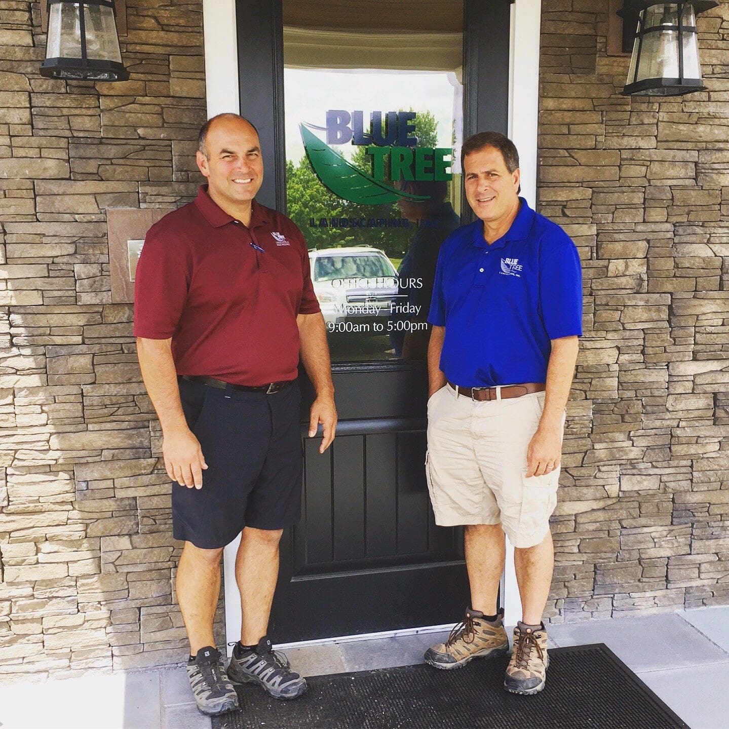 Two men stand smiling in front of a door with a Blue Tree logo. Both wear polo shirts and shorts; one in maroon, the other in blue. The building exterior is stone, and lanterns are mounted on either side of the door.