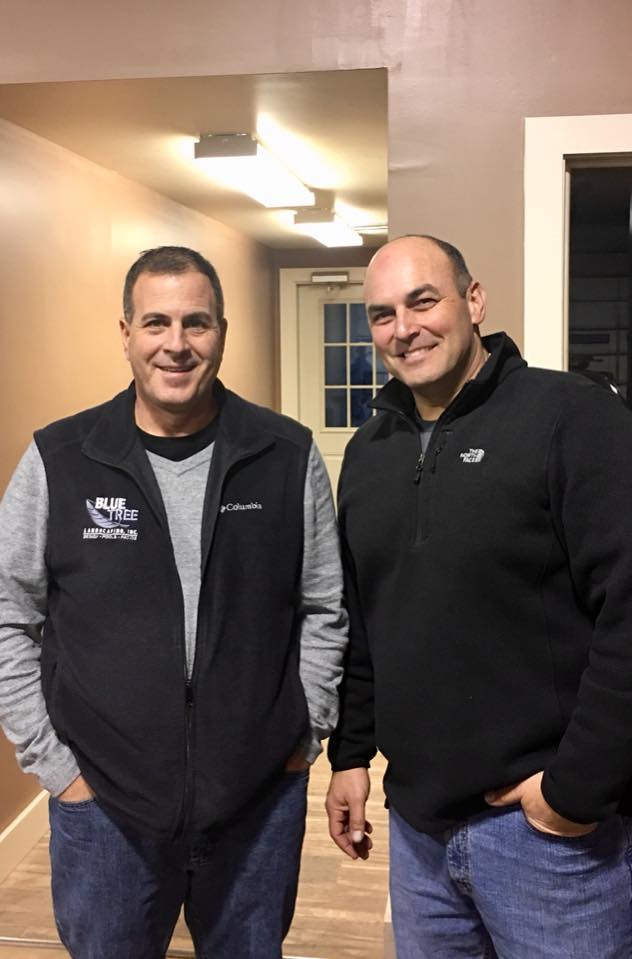 Two men stand indoors, smiling at the camera. Both wear black zip-up jackets and jeans. The man on the left has his hands by his sides, while the man on the right has one hand in his pocket. They are in a hallway with neutral-colored walls.