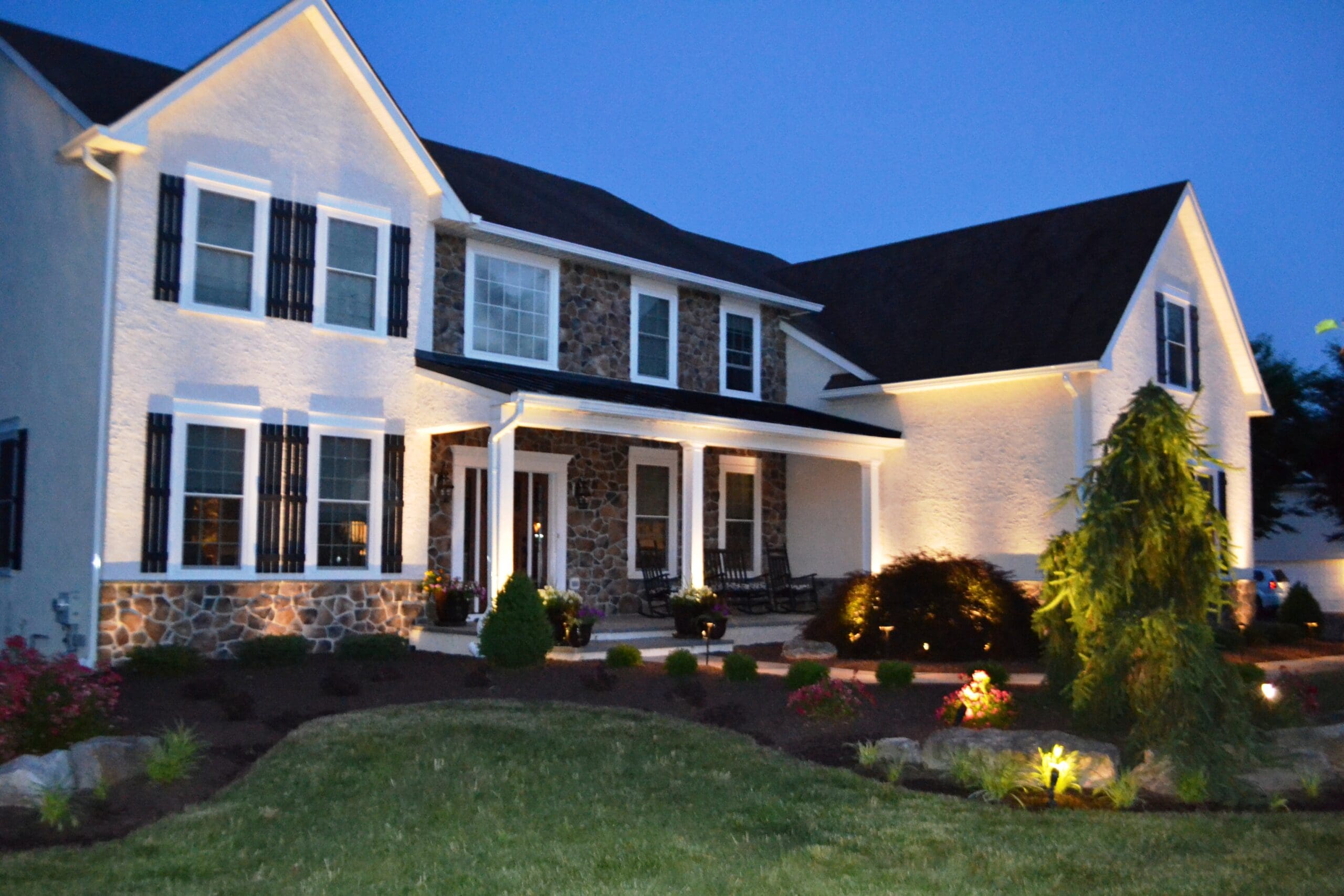 A two-story suburban house with white walls, dark shutters, and stone accents is lit up at dusk. The yard features neatly trimmed bushes, small trees, landscaped flower beds, and outdoor lighting.