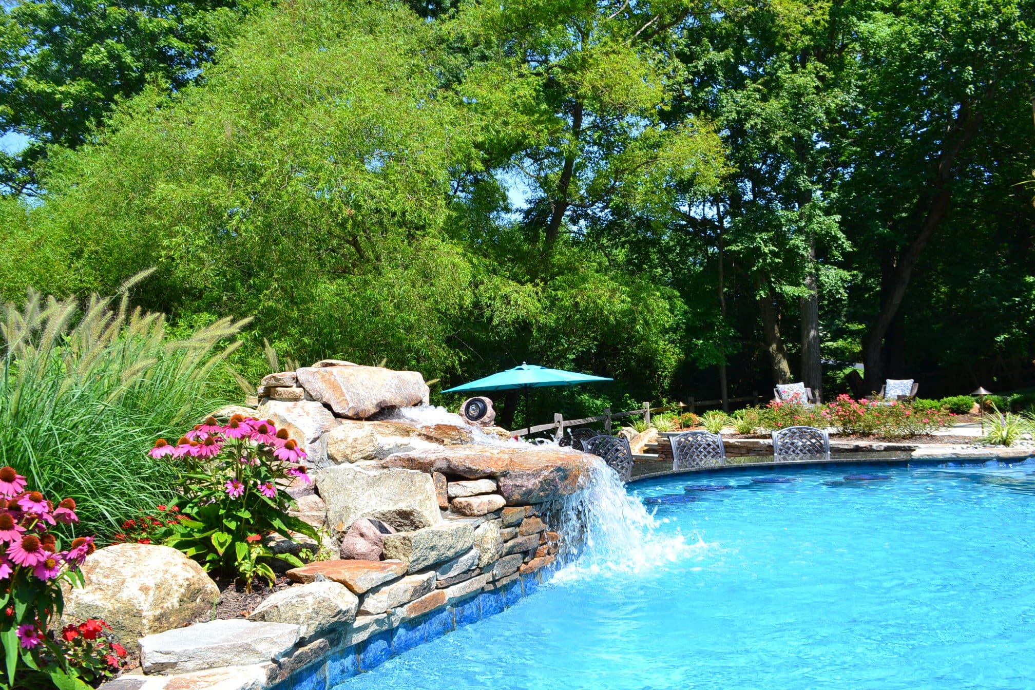 A backyard swimming pool with a stone waterfall feature, surrounded by lush green trees, flowering plants, and a turquoise umbrella providing shade. Lounge chairs are visible in the background.