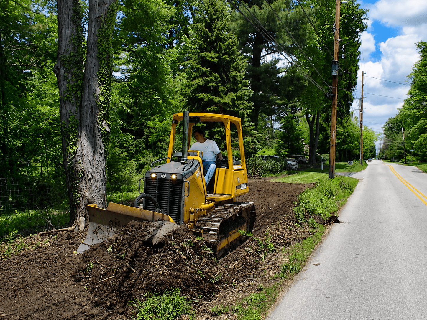 A person operates a yellow bulldozer, clearing soil near a tree along a rural roadside lined with greenery and trees on a sunny day.