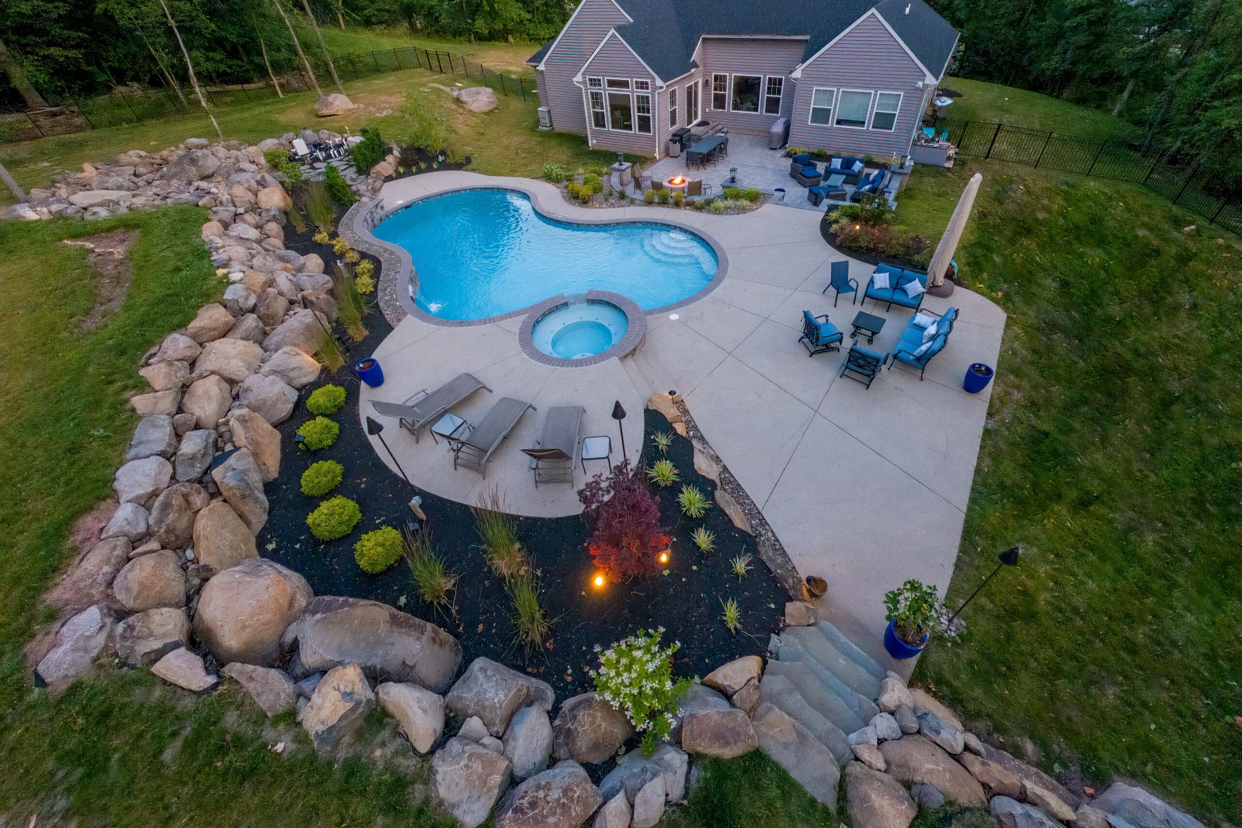 Aerial view of a backyard with a freeform swimming pool, attached hot tub, lounge chairs, patio seating areas, landscaped garden with rocks and plants, and a house in the background.