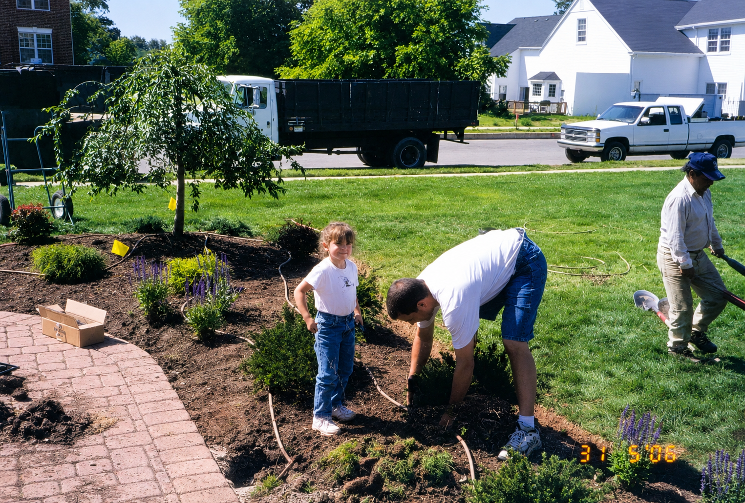 A young girl and an adult are gardening in a yard, with another worker nearby. They are surrounded by bushes and soil, and two pickup trucks are parked on the street in a suburban neighborhood.
