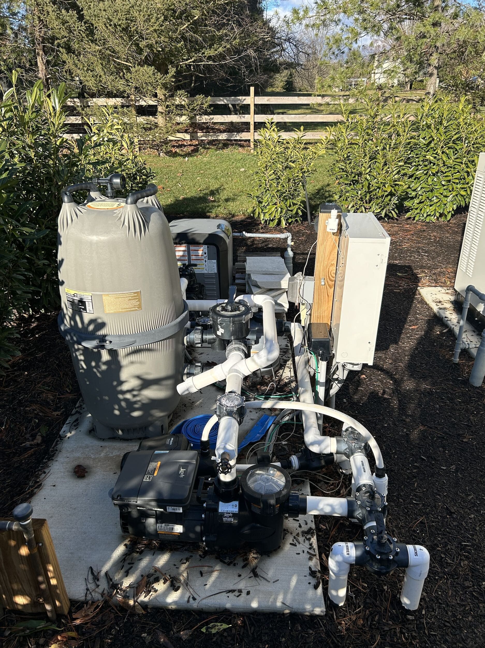 Outdoor pool equipment setup with a filter, pump, electrical box, and various white PVC pipes on a concrete pad, surrounded by mulch, bushes, and a wooden fence in the background under sunlight.