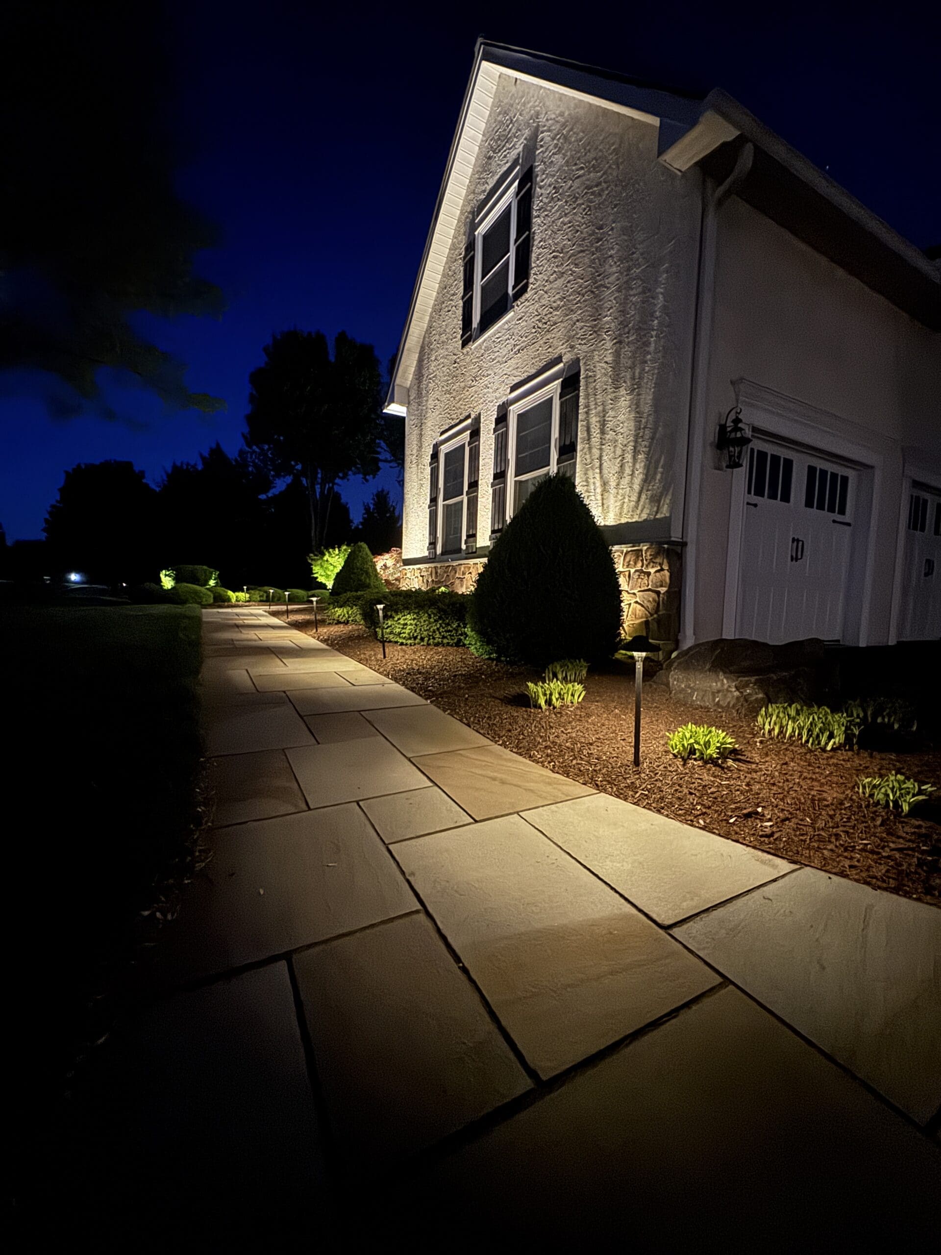 A house at night with exterior lights illuminating a stone walkway, manicured bushes, and garden beds along the side of the building.
