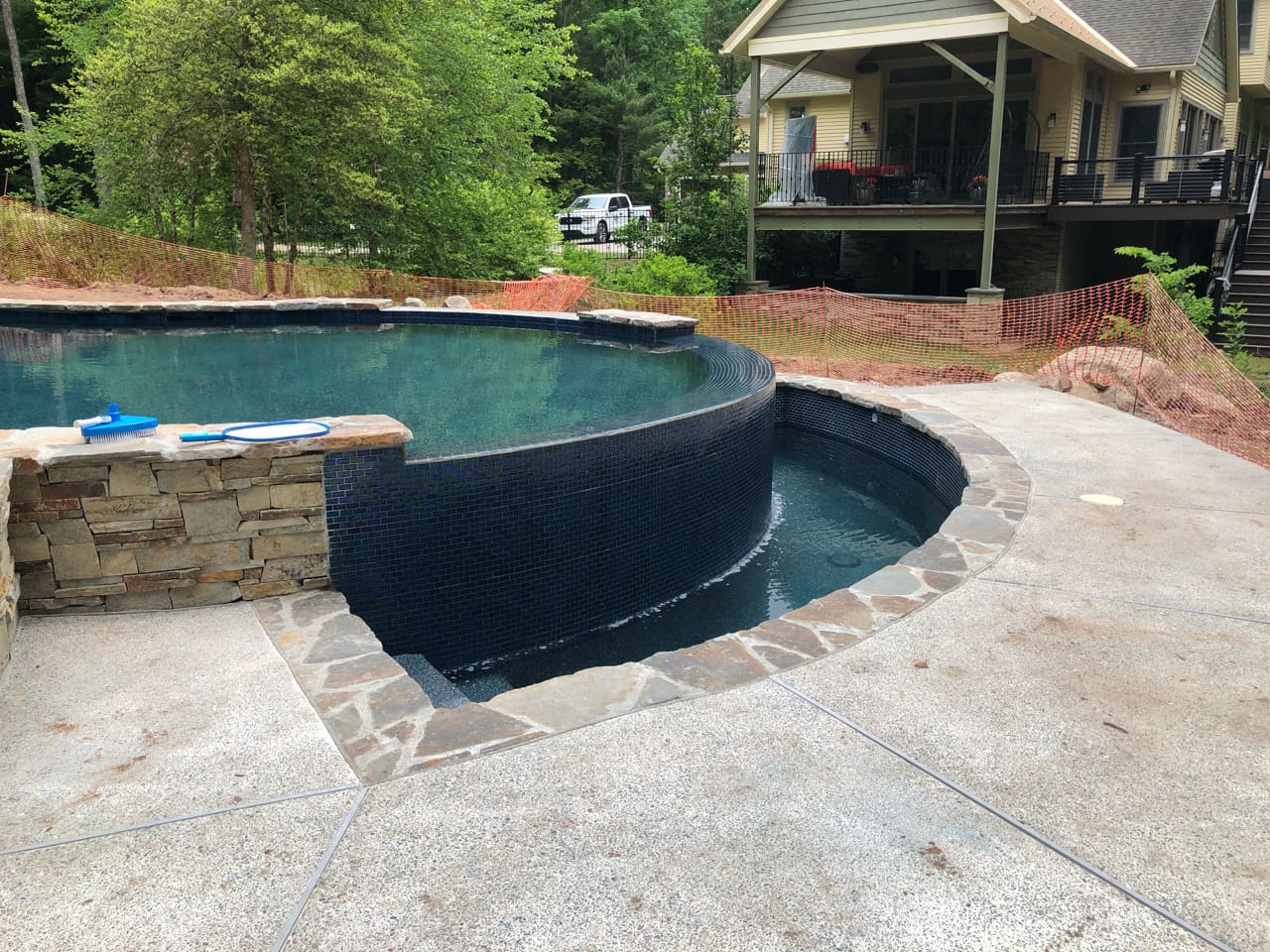 A backyard with a stone infinity-edge pool and a small round lower pool. A house with a porch is in the background, surrounded by trees and some construction materials.