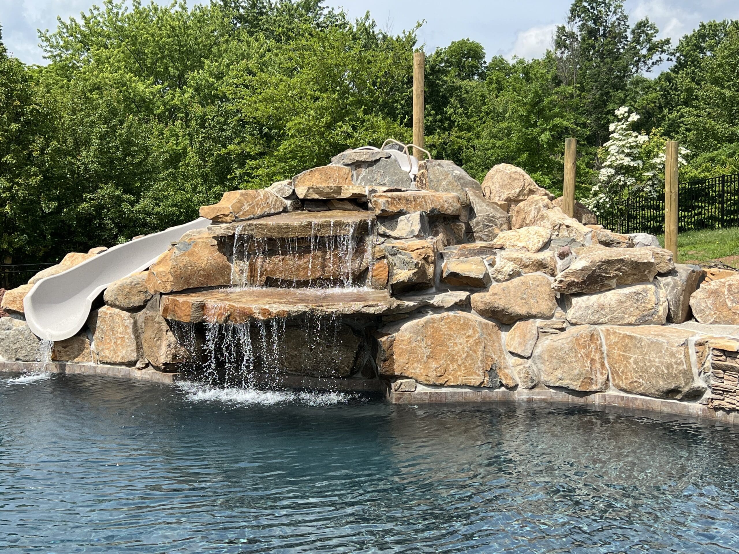 A pool features a stone waterfall and a built-in white slide, surrounded by rocks, with lush green trees in the background under a partly cloudy sky.