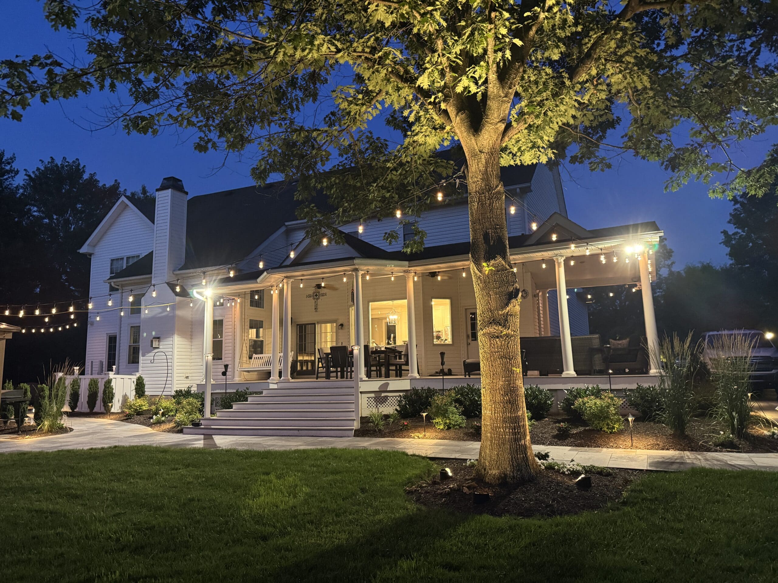 A large white house with a wraparound porch is lit warmly at dusk. String lights hang above the yard, and a tree stands in the foreground, surrounded by neatly landscaped grass and plants.