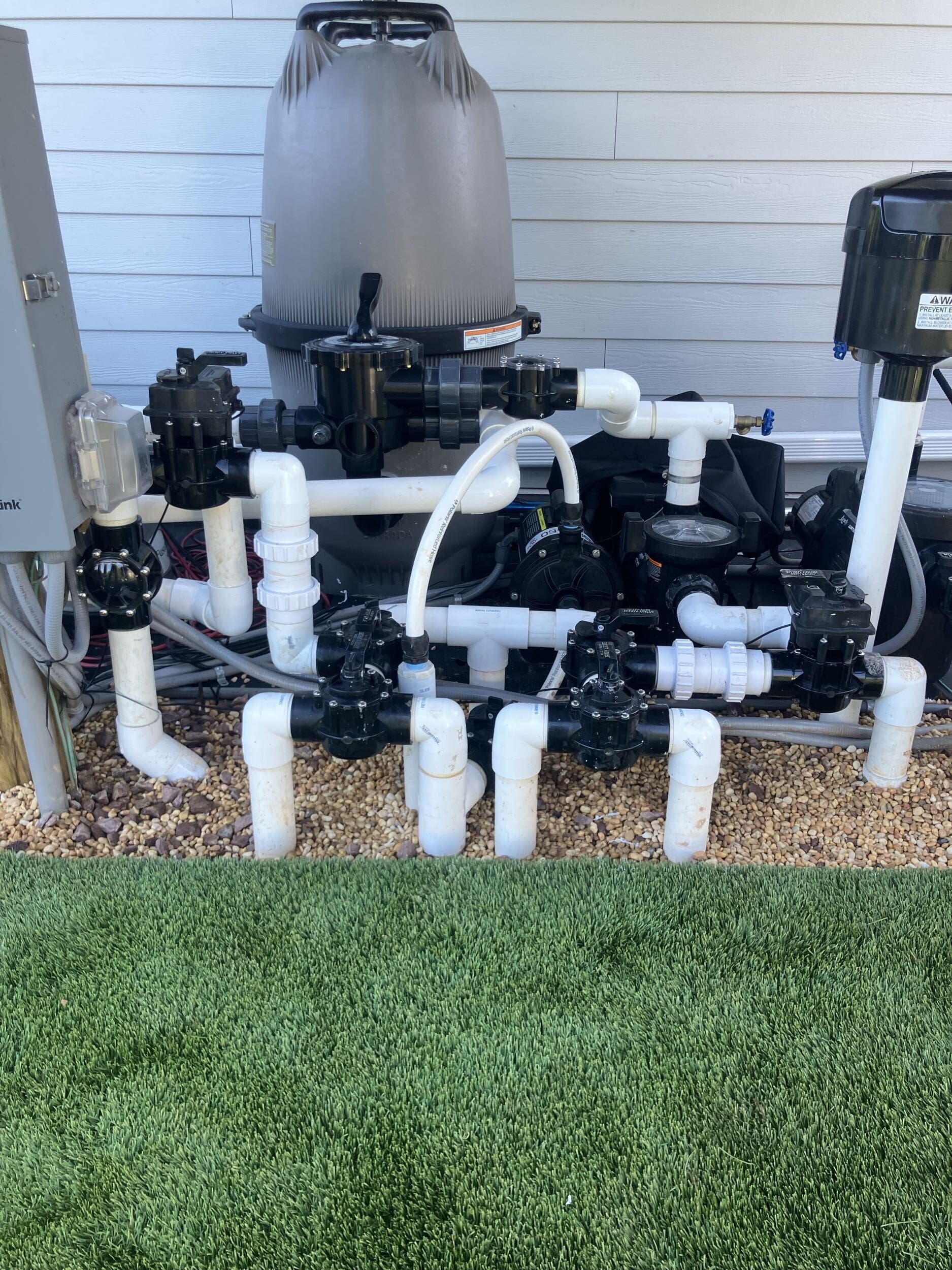 A complex pool pump system with multiple white PVC pipes, black valves, and a large filter is installed on gravel next to artificial grass and a white wall.