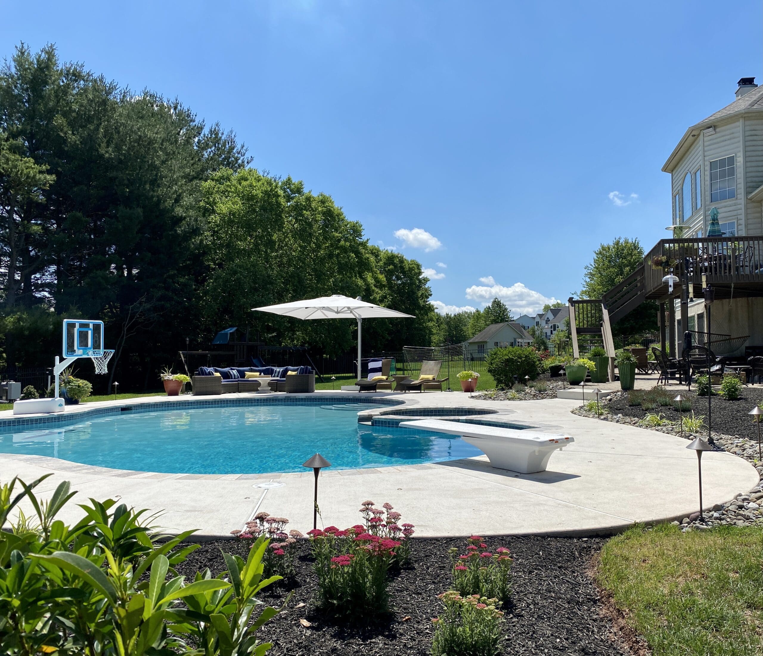 A backyard with a large pool, diving board, basketball hoop, lounge chairs, umbrella, and lush greenery. A house and deck are visible on the right under a bright blue sky.