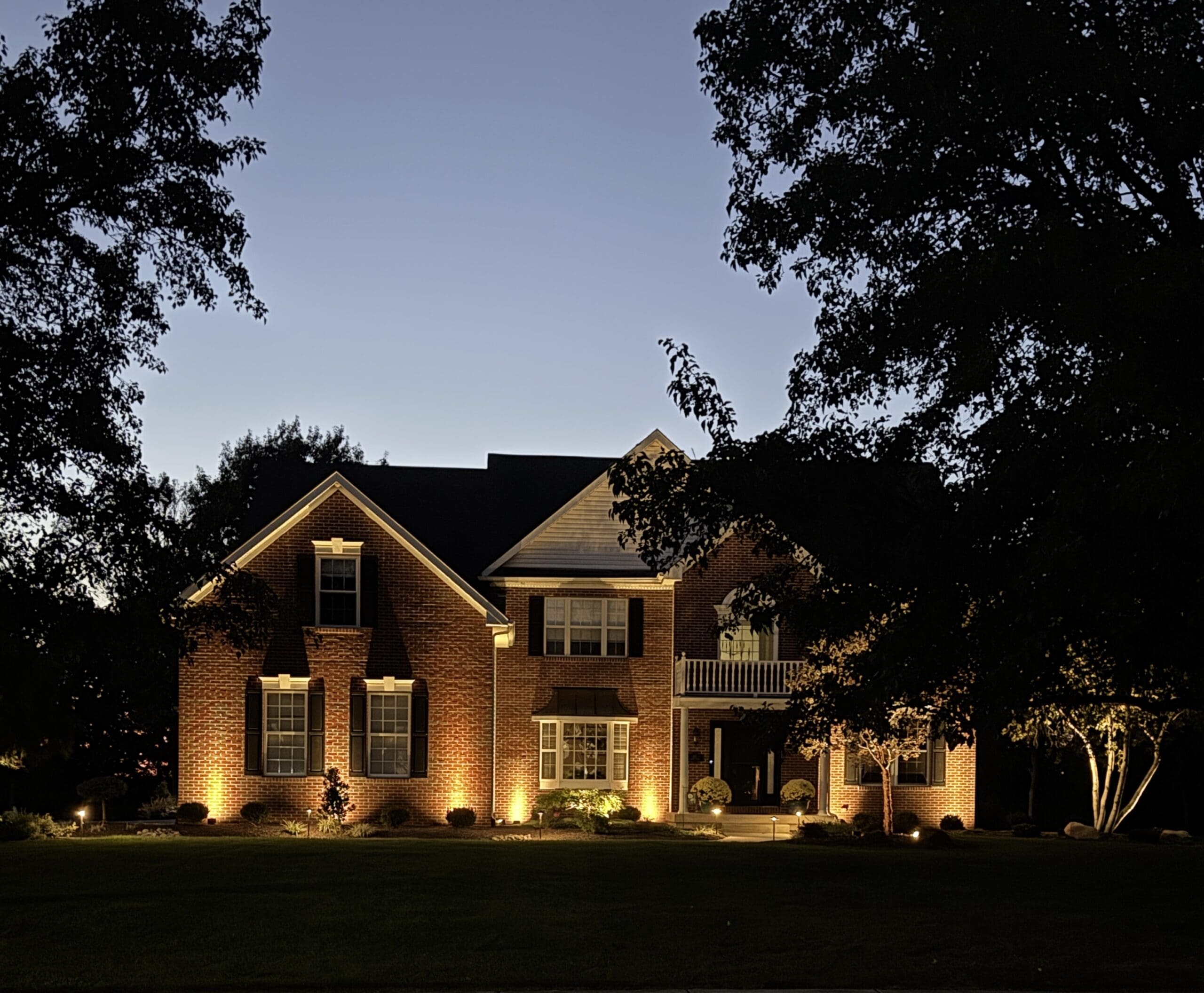 A two-story brick house with exterior lights illuminating the front at dusk, surrounded by trees and a dark lawn, under a clear evening sky.