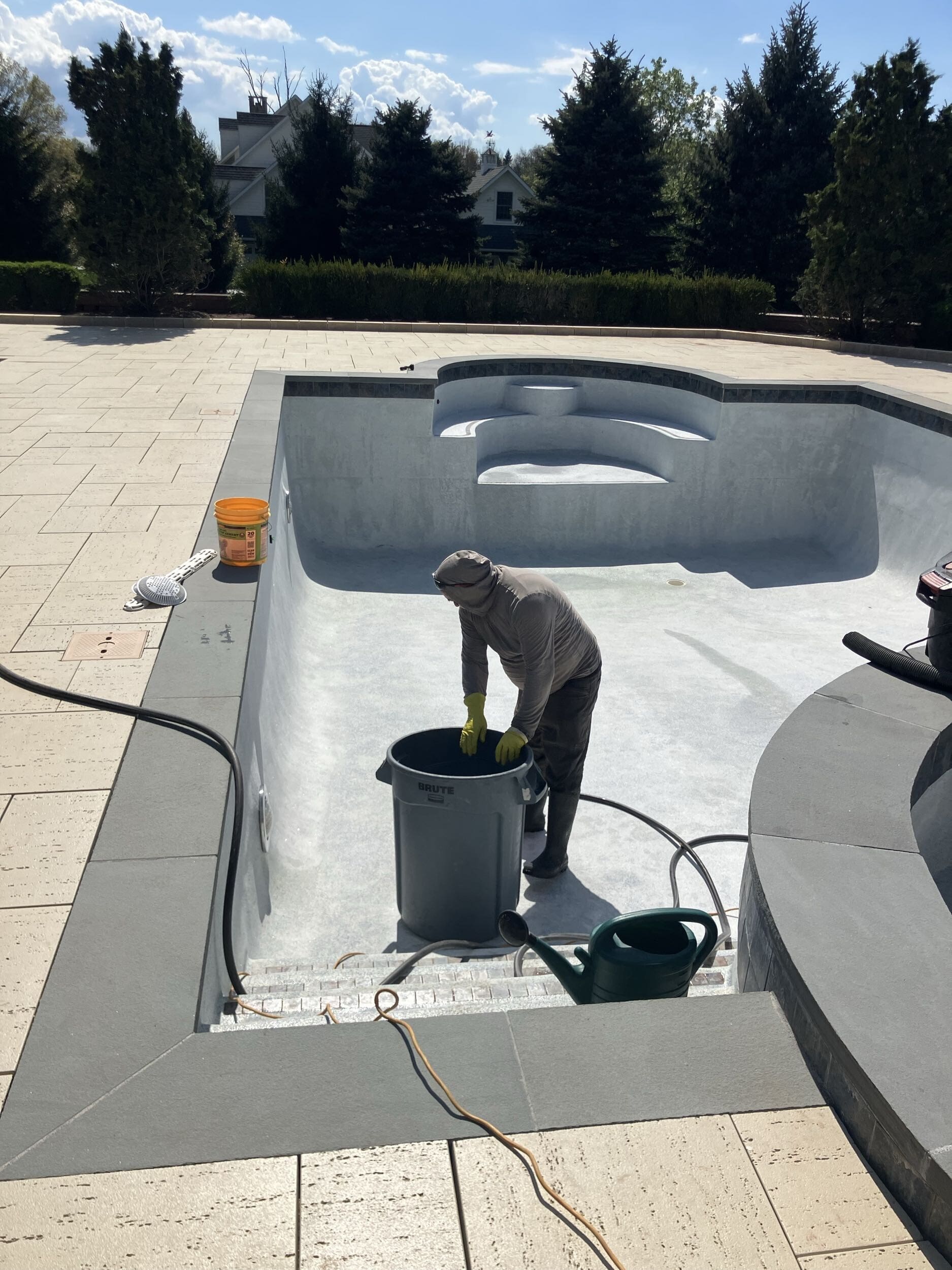 A person wearing protective clothing and gloves stands inside an empty swimming pool, cleaning with a trash bin nearby. A yellow bucket, garden hose, and vacuum are on the pool’s edge under a sunny sky.