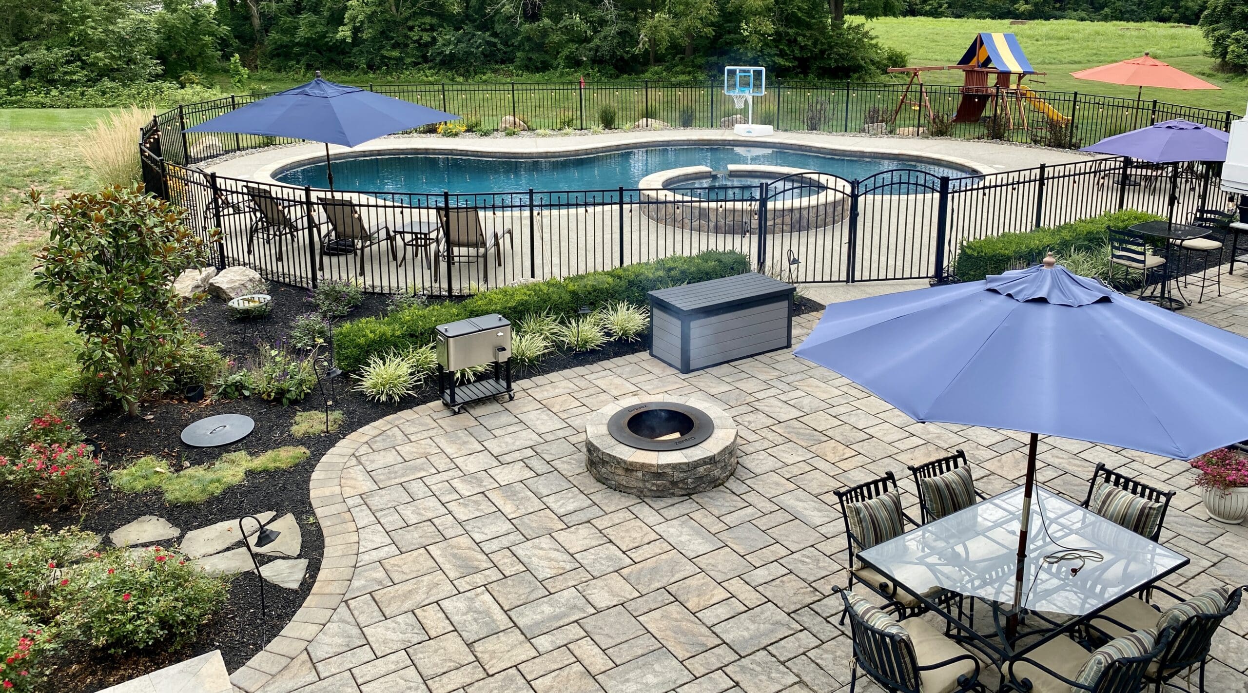 Backyard patio with dining table, umbrella, fire pit, grill, and landscaped plants. A fenced-in pool, basketball hoop, and playground are in the background, surrounded by green grass and trees.