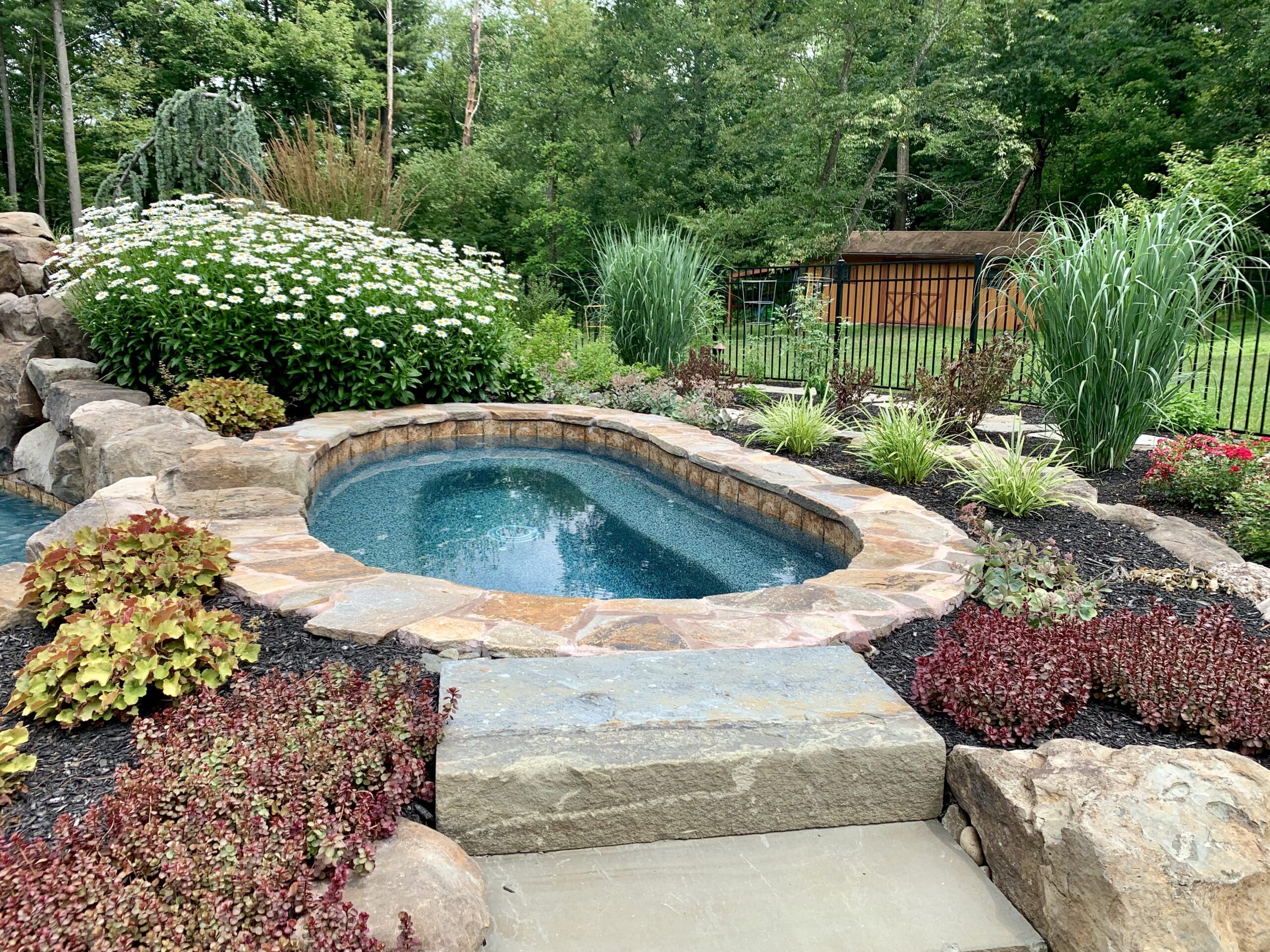 A small, stone-edged hot tub is surrounded by lush greenery, flowering plants, and ornamental grasses in a landscaped backyard with a wooden shed and trees in the background.