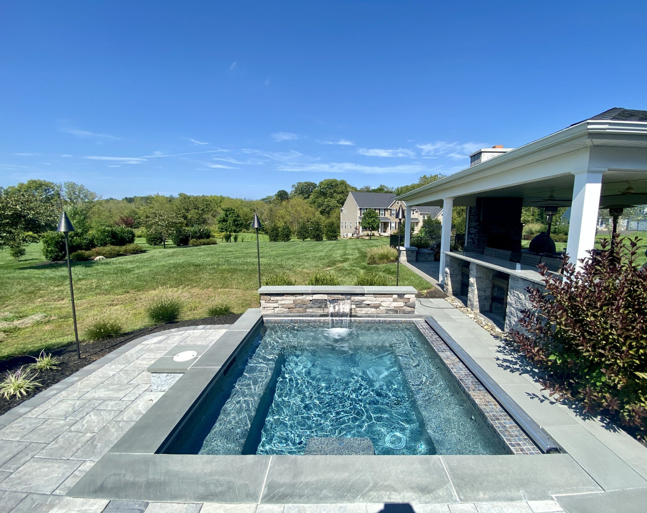 A small, backyard pool with clear water and a stone waterfall feature, surrounded by a landscaped patio and greenery. A covered outdoor seating area is on the right, with a grassy lawn and trees in the background under a clear blue sky.
