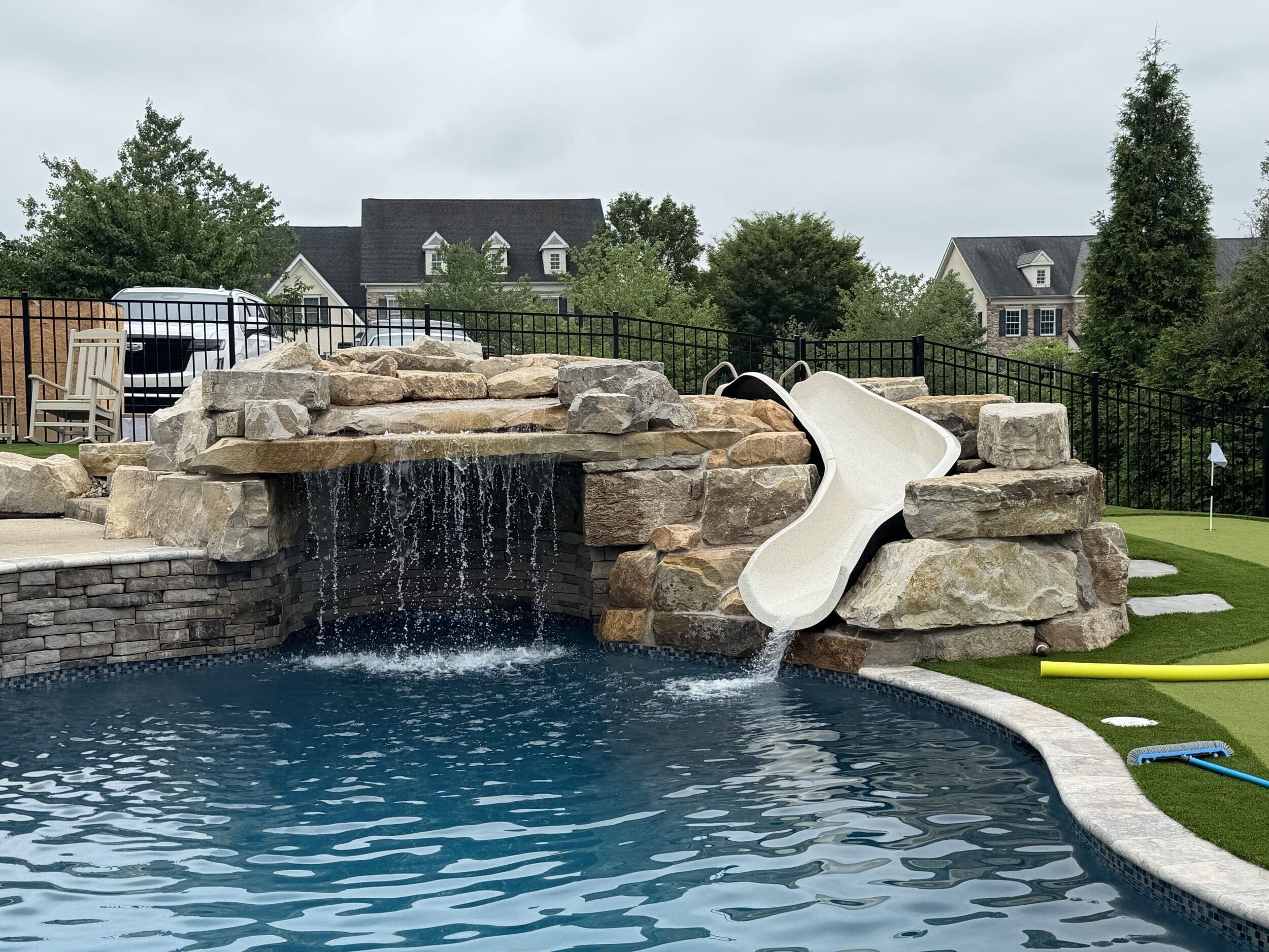 A backyard pool features a rock waterfall and a white slide curving into the water. Surrounding the pool are artificial grass, scattered pool toys, and houses in the background under a cloudy sky.