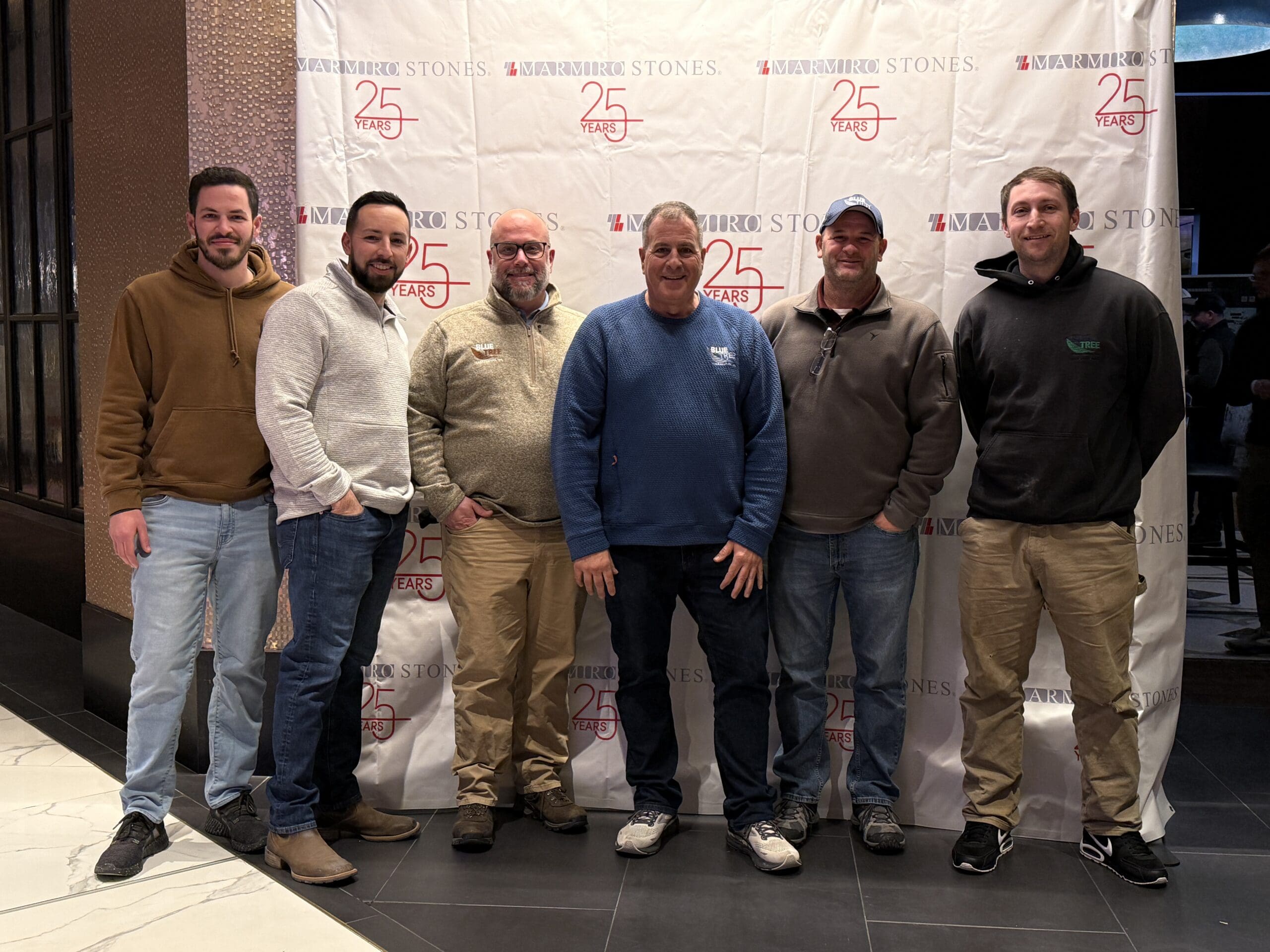 Six men stand in front of a step-and-repeat banner celebrating 25 Years of Warwick Stones. They are smiling and dressed casually in sweaters, hoodies, and jackets, posing indoors on a tiled floor.