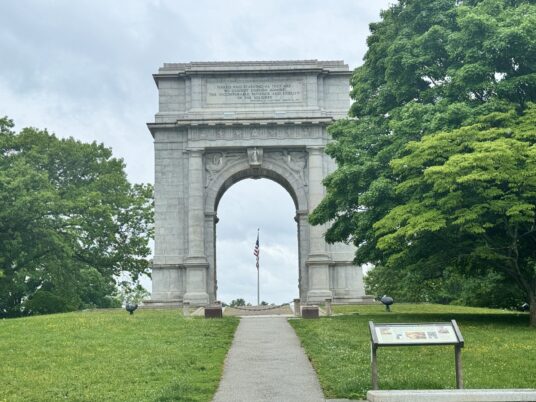 A large stone triumphal arch stands on a grassy hill, flanked by green trees. An American flag is visible through the arch, and an informational sign is in the foreground beside a paved path.