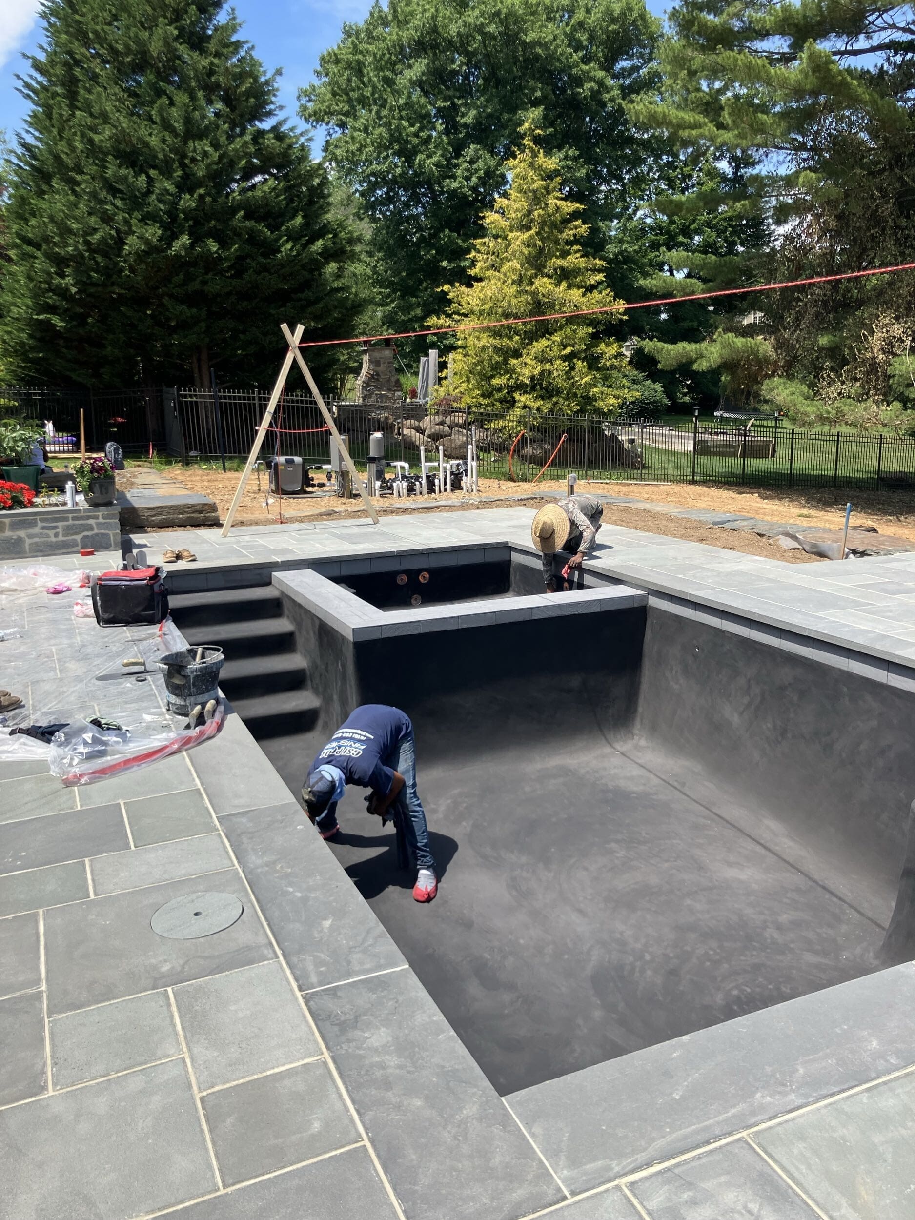 Two workers are constructing or finishing a rectangular in-ground pool surrounded by stone tiles, with equipment and materials nearby, and trees and a black fence in the background.