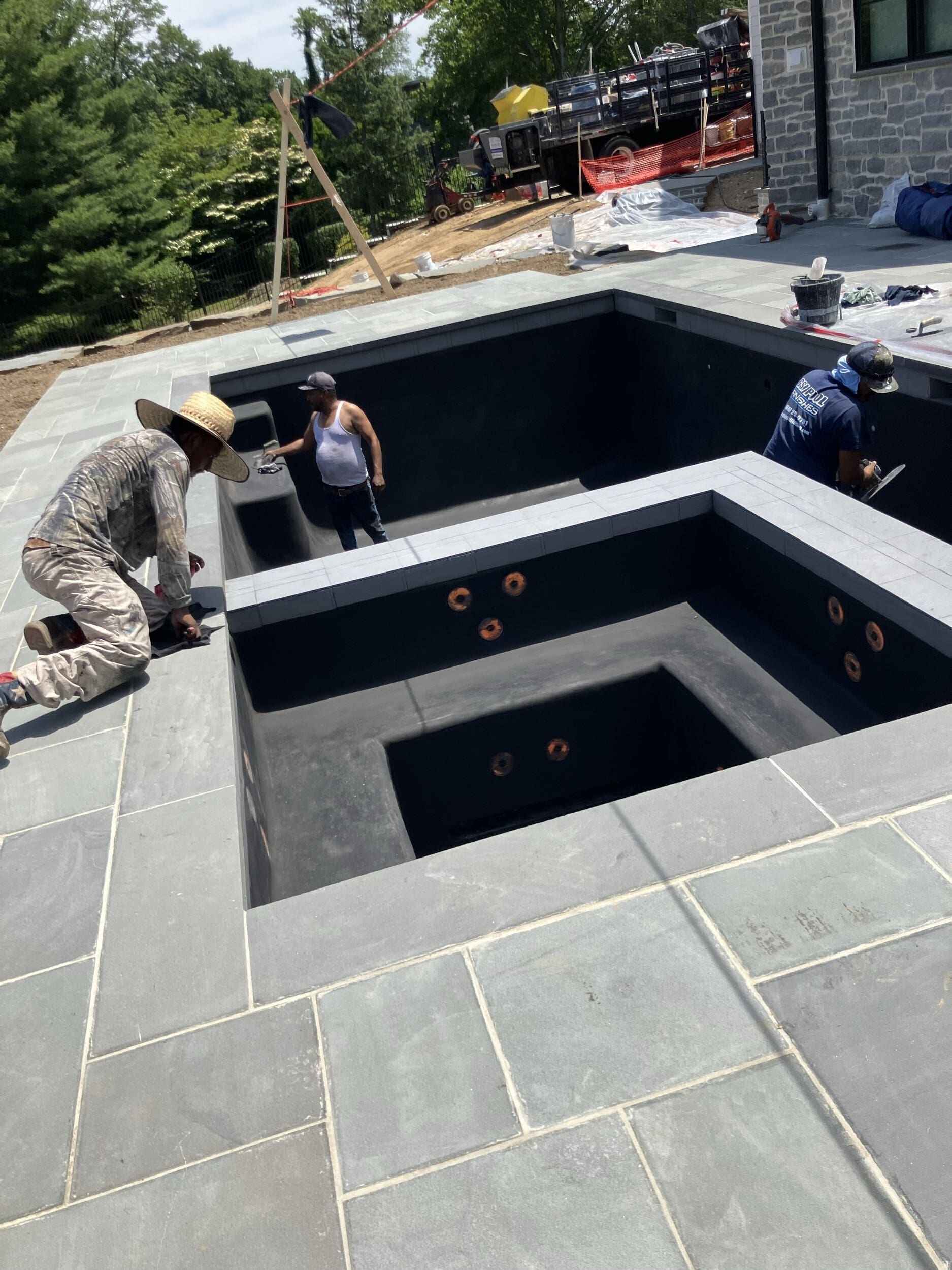 Three workers in hats and work clothes install or tile the interior of an empty, unfinished swimming pool on a sunny day, surrounded by gray stone patio tiles and construction materials.