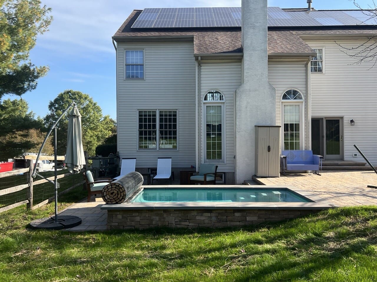 A backyard with a small rectangular pool, lounge chairs, a closed patio umbrella, and a light-colored two-story house with solar panels on the roof in the background.