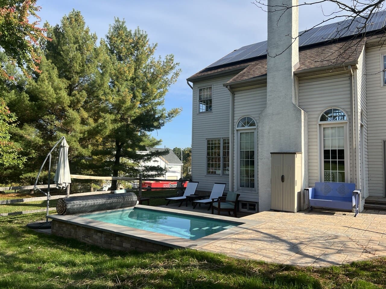 A small backyard pool beside a beige house with tall windows and a stone patio, surrounded by trees and outdoor furniture, including lounge chairs and a blue bench.