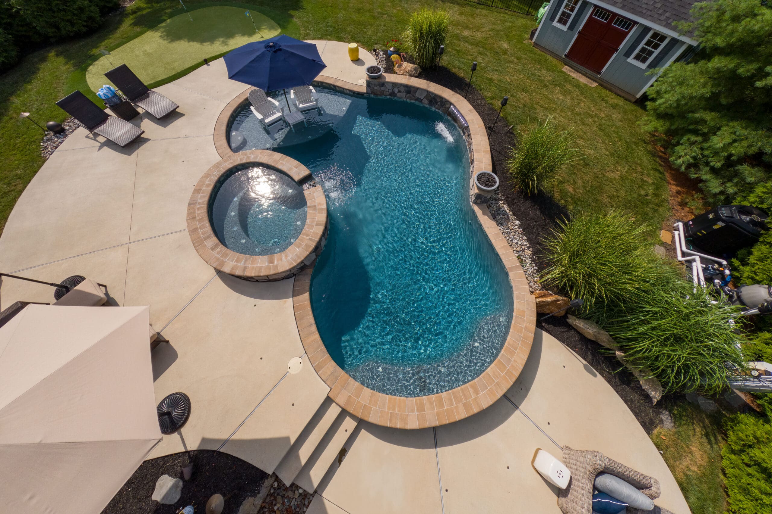 Aerial view of a backyard with a kidney-shaped pool, attached spa, lounge chairs, umbrellas, landscaped greenery, and a small shed in one corner.