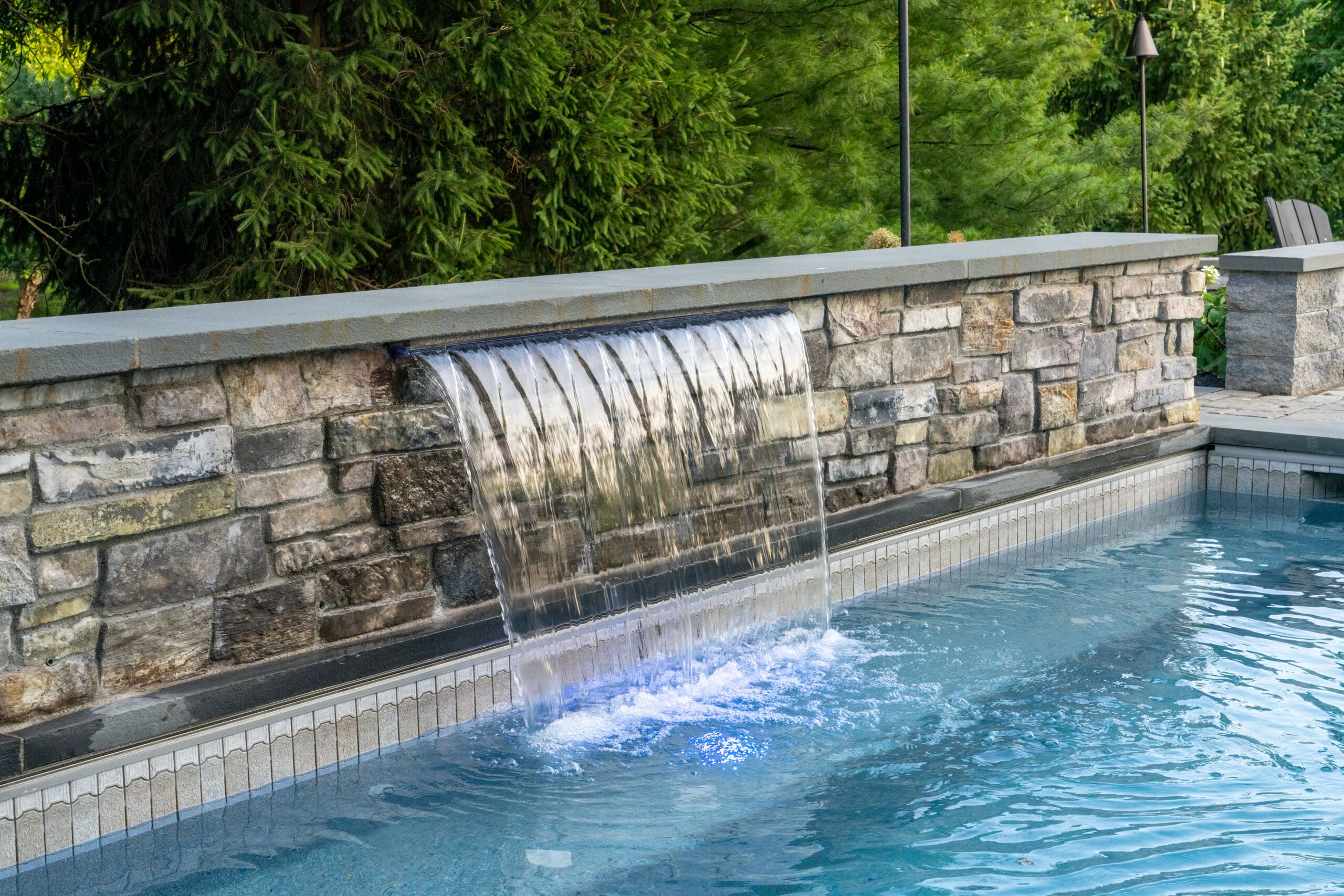 A stone wall with a built-in waterfall feature pours water into a clear swimming pool, surrounded by lush green trees and outdoor seating.