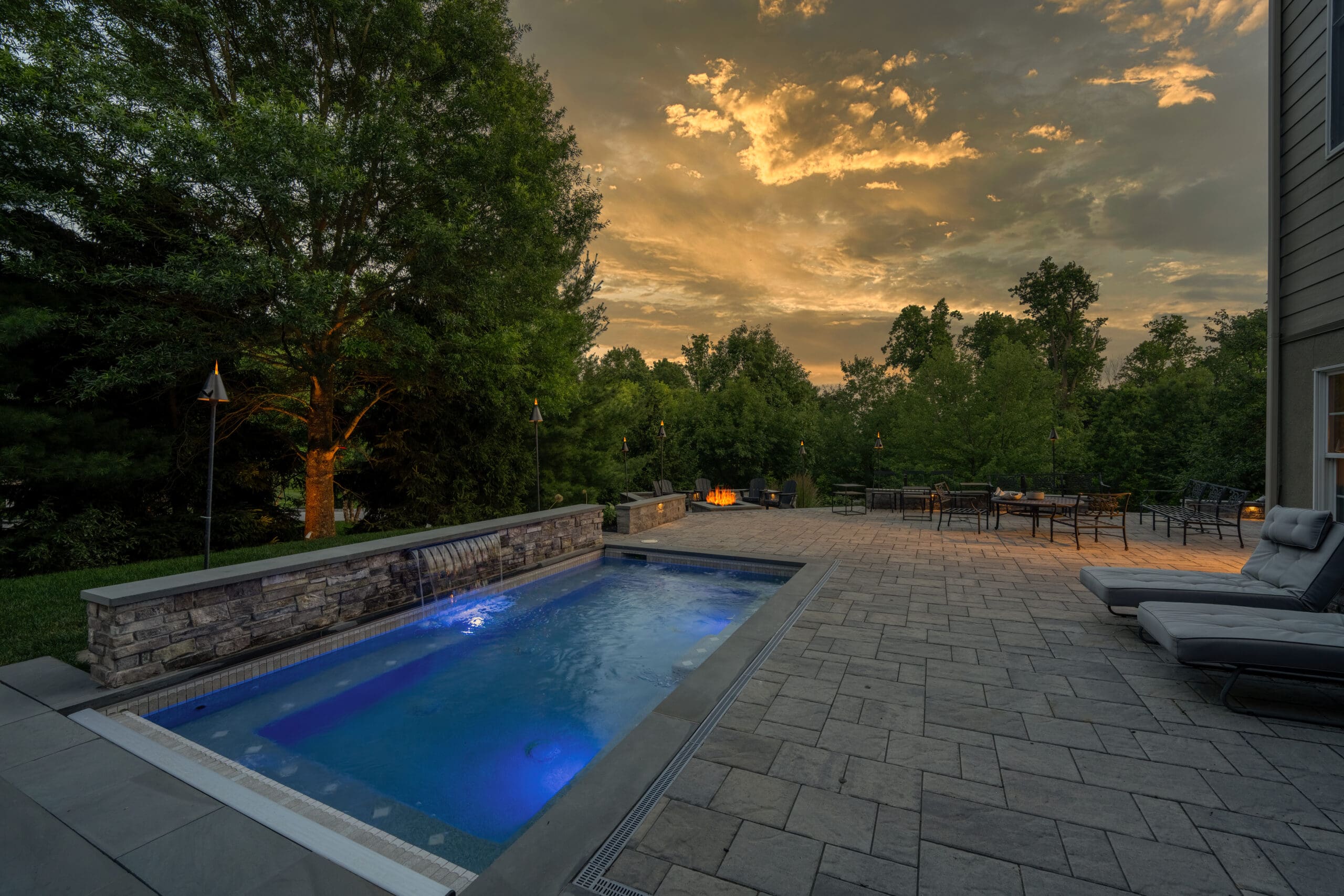 A modern backyard features a small, illuminated pool with blue lights, surrounded by stone pavement, lounge chairs, a dining area, lush trees, and a dramatic sunset sky.