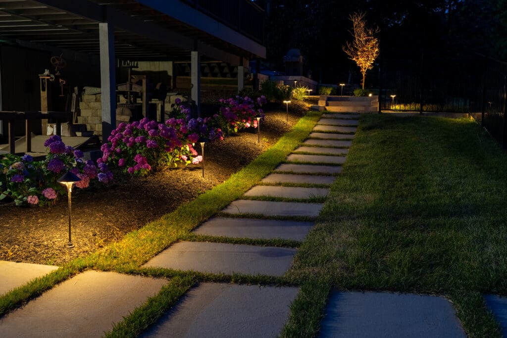 Landscape lights illuminate a garden path, flowering plants, and a tree beside an outdoor living space at night.