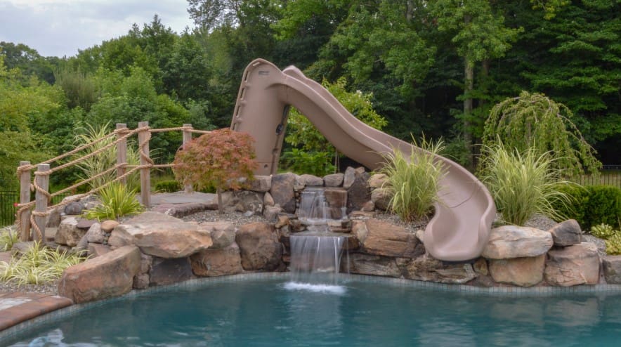 A curved brown water slide built over rocks leads into a swimming pool, with a small waterfall flowing beneath the slide. Lush green trees and plants surround the pool area.