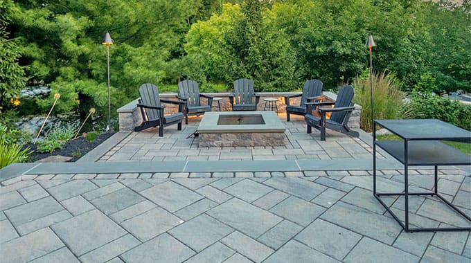 Patio with gray stone pavers, a square fire pit in the center, five Adirondack chairs arranged around it, torches, and lush green trees and plants in the background.