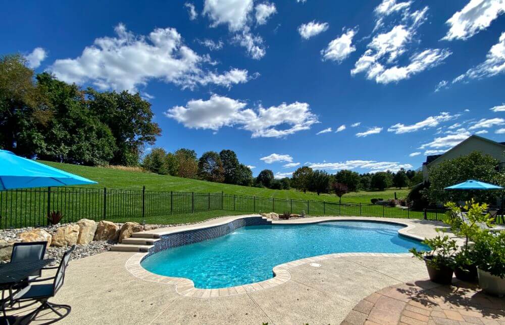 A clear swimming pool with curved edges sits beside a green lawn and trees under a bright blue sky with scattered clouds. Patio furniture and blue umbrellas are set on the concrete pool deck.