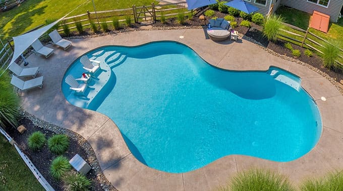 Aerial view of a backyard with a curvy, in-ground swimming pool, lounge chairs partially submerged in the water, patio chairs under umbrellas, and landscaping with grass and shrubs around the pool.