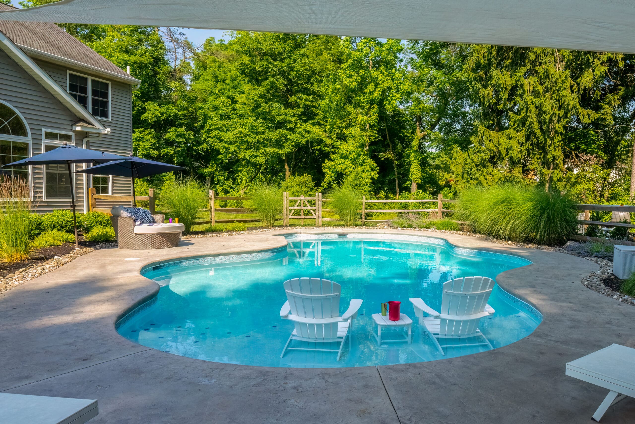 A backyard pool with two white Adirondack chairs and a small table in the shallow end, surrounded by greenery, a wooden fence, and patio furniture under a blue umbrella.