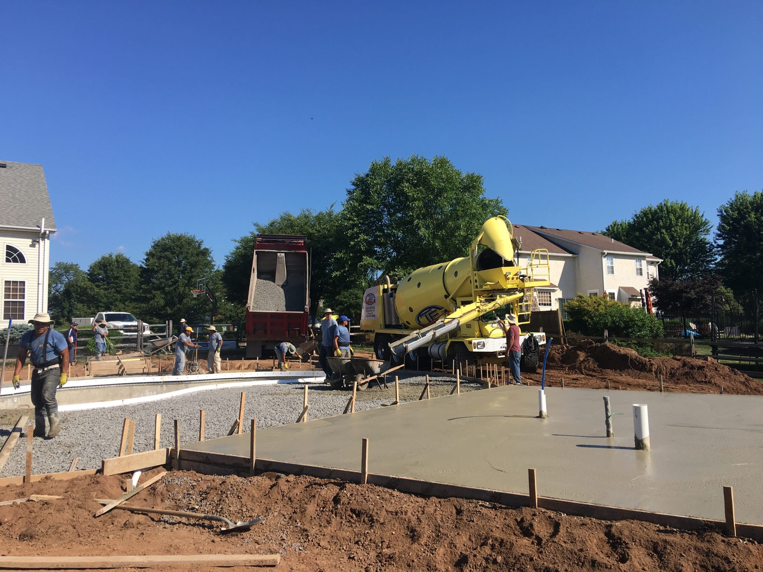 Construction workers pour concrete from a yellow cement truck onto a residential building foundation, surrounded by wooden framing and houses on a sunny day.