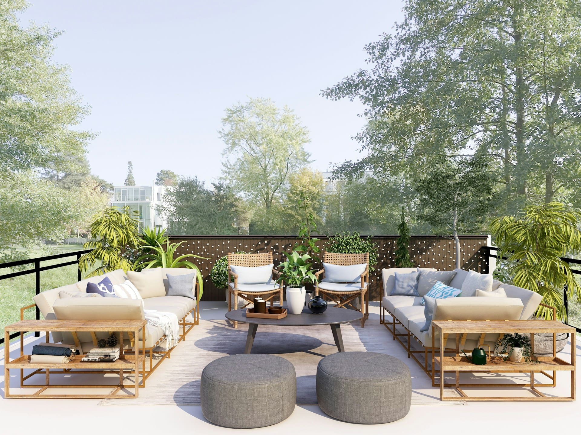 A modern outdoor patio with two sofas, two armchairs, a round coffee table, two gray ottomans, potted plants, and decorative pillows, set on a wooden deck surrounded by trees and greenery.