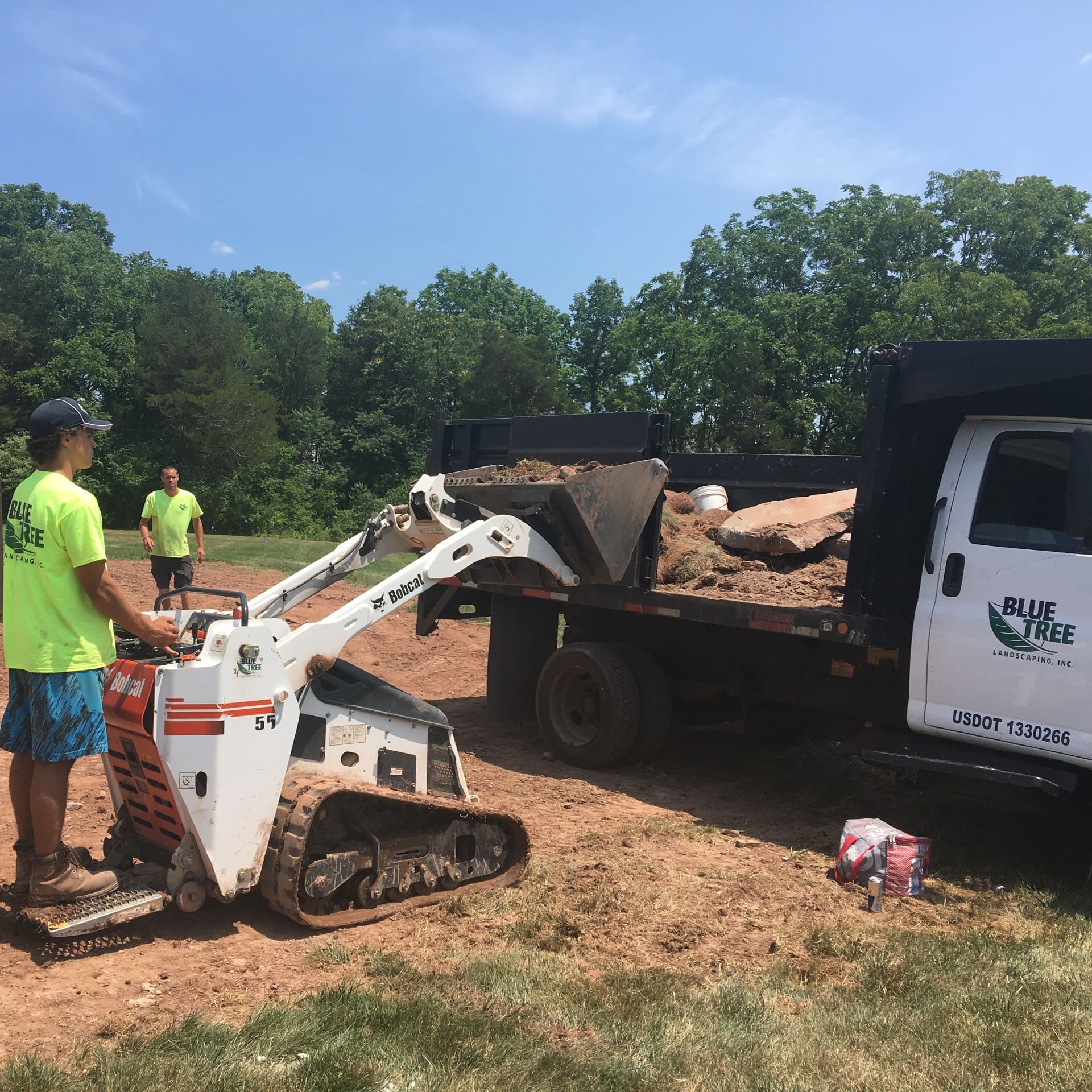 A worker operates a small loader to dump soil and debris into a Blue Tree Landscaping truck, with another worker standing nearby on a sunny day outdoors.