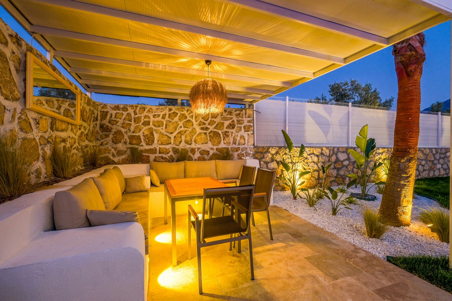 A cozy outdoor patio with cushioned seating, a wooden table, and chairs under a covered pergola. Warm lighting highlights stone walls, plants, and a hammock, creating an inviting evening atmosphere.