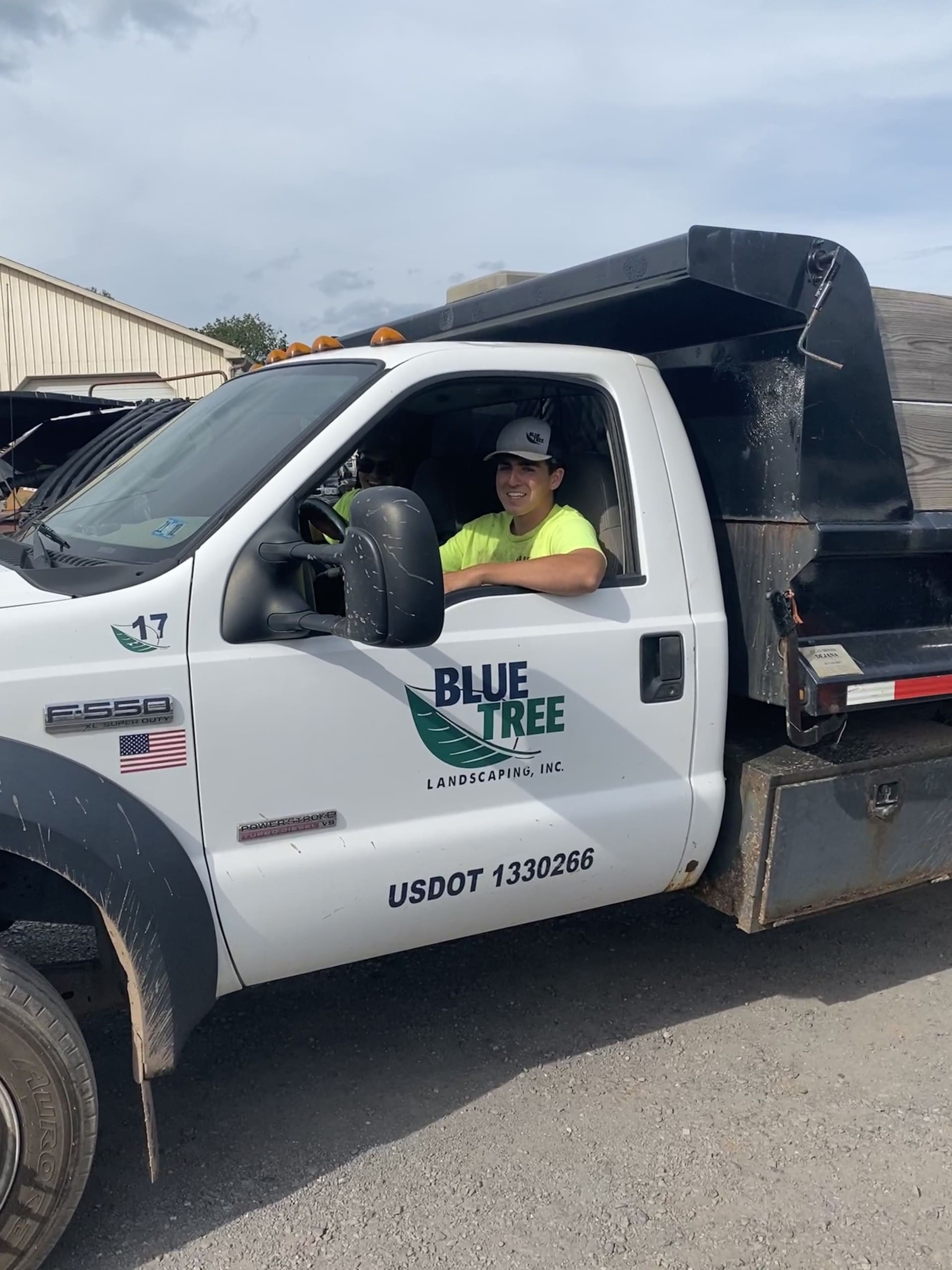 A person wearing a yellow shirt and cap smiles while sitting in the driver’s seat of a white Blue Tree Landscaping Inc. truck parked beside a building. The door displays the company logo and USDOT number.