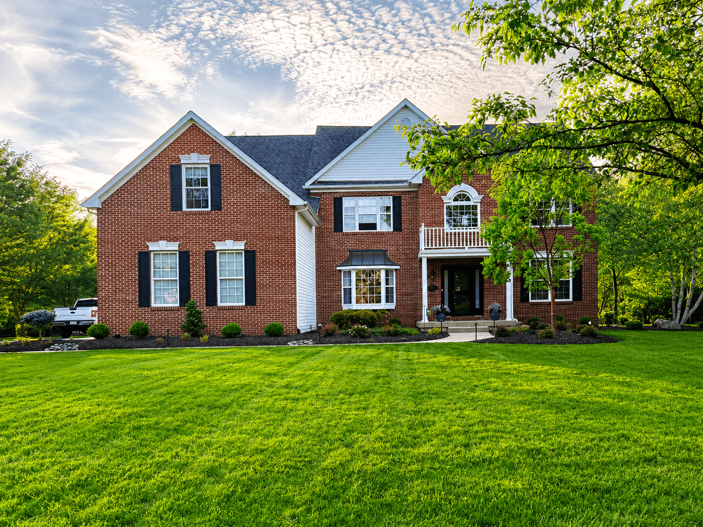 A large brick two-story house with white trim, black shutters, and a neatly manicured lawn sits beneath a partly cloudy sky, surrounded by green trees and shrubs.