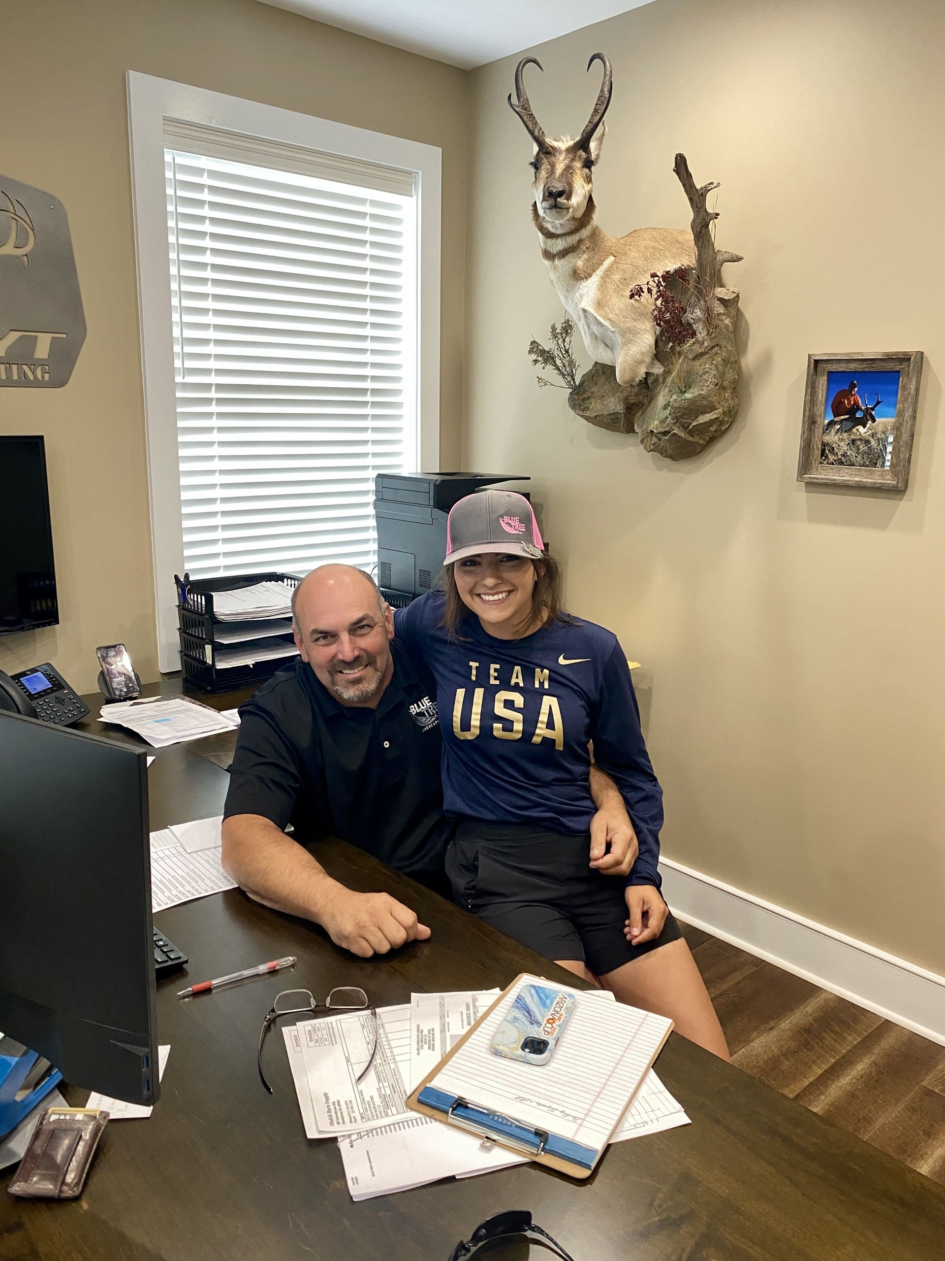 A smiling man sits at a desk with paperwork while a woman in a Team USA shirt and hat sits on his lap. Behind them are a mounted antelope head and framed photos on a beige wall.