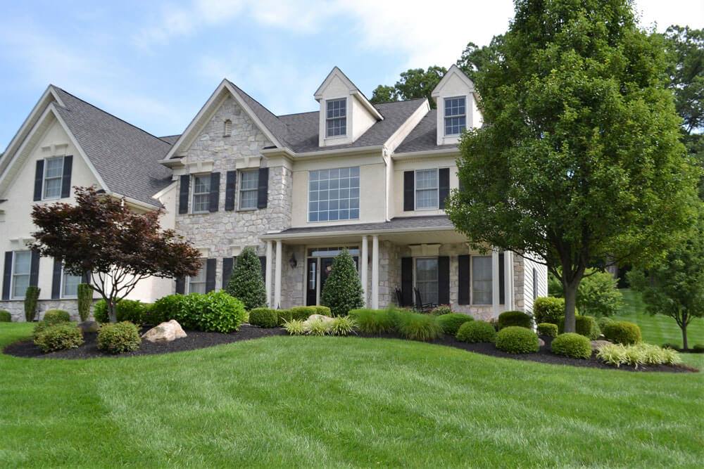 A large, two-story house with stone and beige siding, black shutters, and multiple gables. The front yard features manicured shrubs, green grass, and several trees under a blue sky.
