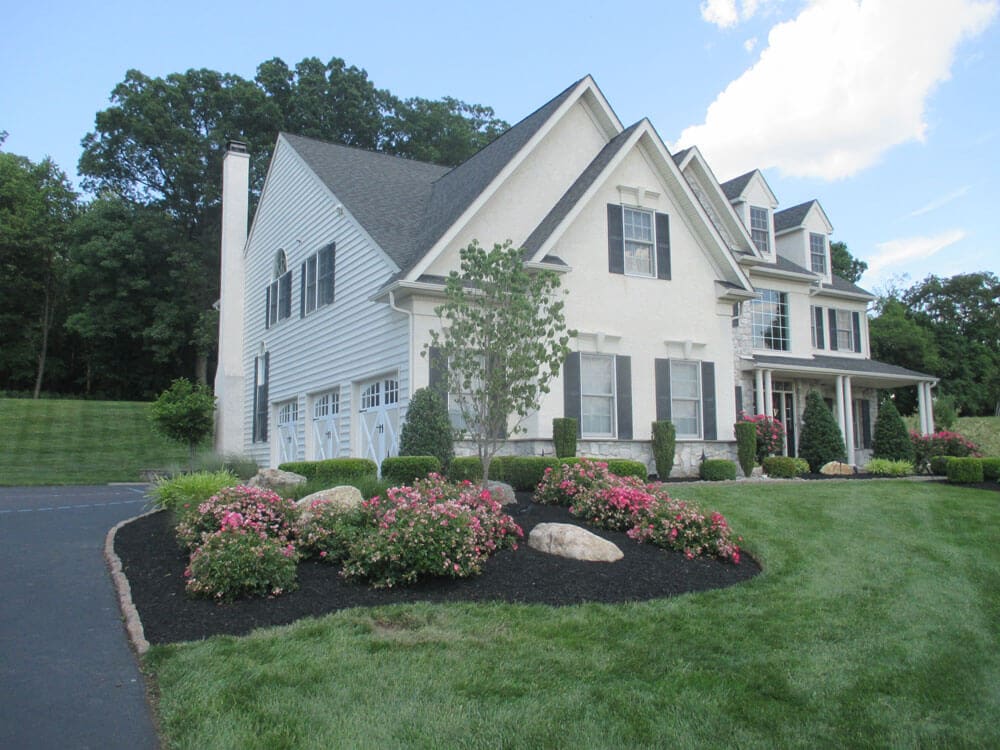 A large two-story suburban house with white siding and black shutters, surrounded by neatly trimmed grass, landscaped flower beds with pink flowers, and a driveway on the left. Trees are visible in the background.