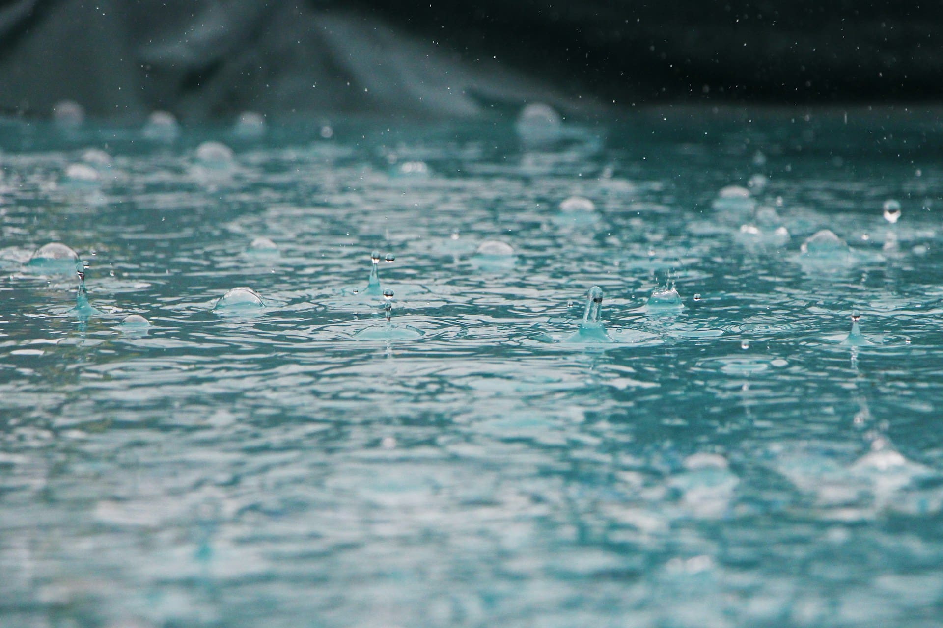 Close-up of raindrops splashing and creating ripples on the surface of turquoise water, with blurred background and droplets frozen mid-air.