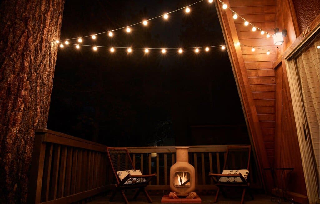 A cozy patio at night features two wooden chairs with pillows, a small outdoor fireplace with a fire burning, and string lights hanging overhead, all next to a wooden cabin.