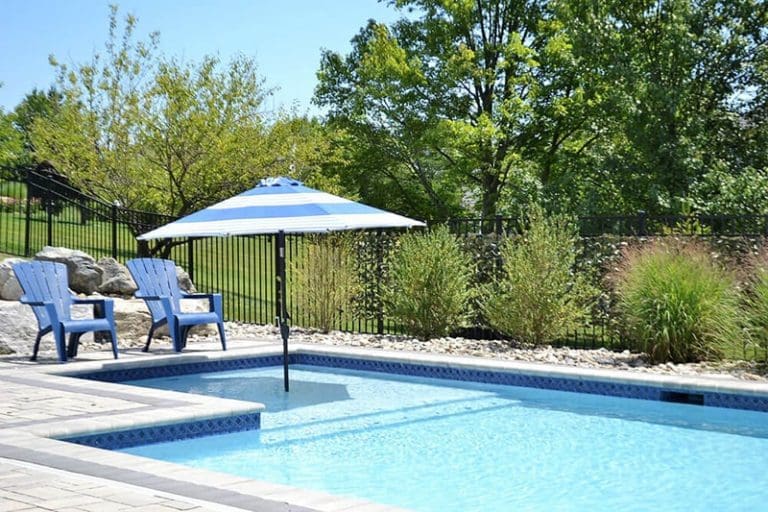 A swimming pool with clear blue water, two blue Adirondack chairs, and a blue-and-white striped umbrella. Lush green trees and plants surround the pool, and there is a black metal fence in the background.