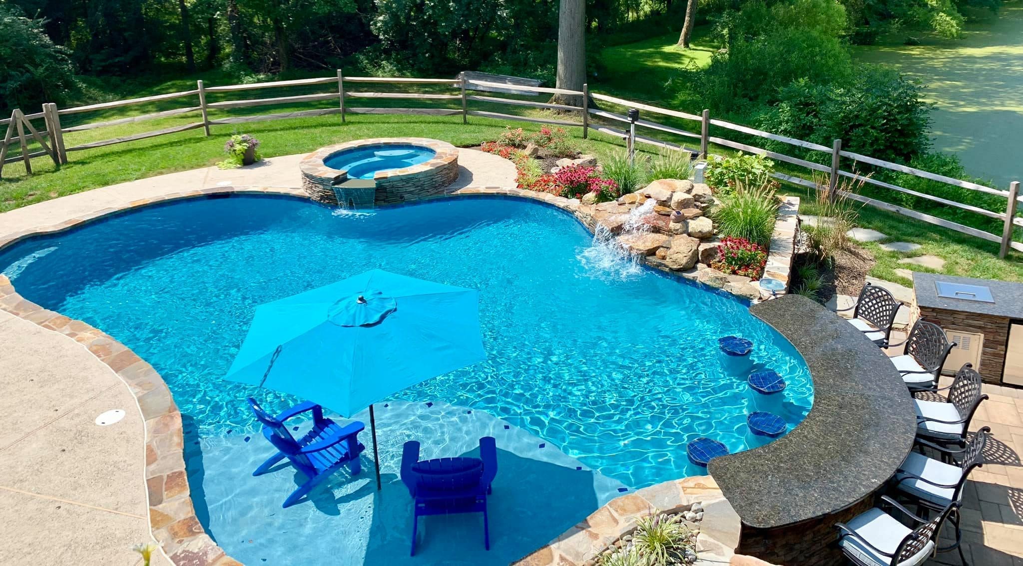 A backyard pool with a hot tub, waterfall, swim-up bar with stools, blue chairs and umbrella in the shallow end, surrounded by landscaped gardens, lawn, and a wooden fence near a pond.