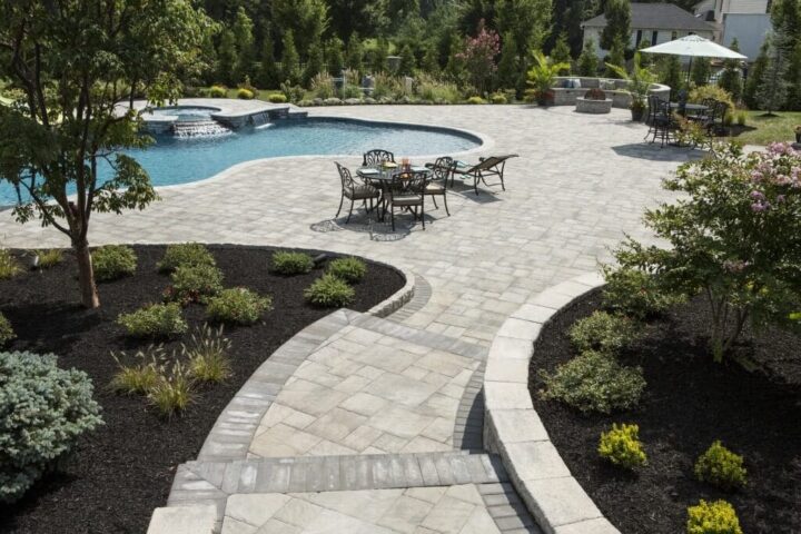A landscaped backyard features a stone patio with a curved walkway, outdoor dining table and chairs, and a swimming pool surrounded by greenery and trees. Manicured garden beds line the paved area.